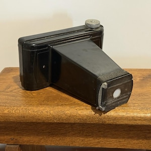 Könnte beinhalten: Eine schwarze Vintage-Kamera mit rechteckigem Gehäuse und einem hervorstehenden Objektivteil. Die Kamera befindet sich auf einer Holzoberfläche und zeigt ihr klassisches Design und ihr gealtertes Aussehen. Die Oberseite hat ein silberfarbenes Zifferblatt.