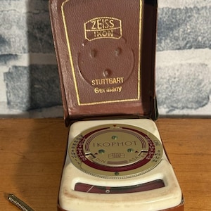 Könnte beinhalten: Vintage Zeiss Ikon Ikophot Belichtungsmesser in einem braunen Lederetui. Das Messgerät hat ein weißes Zifferblatt mit einem kreisförmigen Zifferblatt und roten Akzenten. Das Etui ist mit dem Zeiss Ikon Logo und "Stuttgart Germany" geprägt.