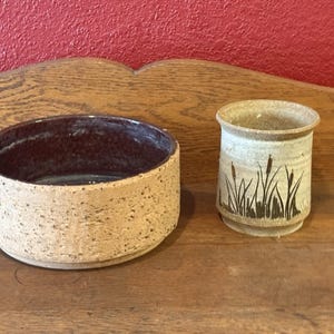 Puede incluir: Dos piezas de cerámica hechas a mano: un cuenco bajo y ancho con un exterior beige moteado y un interior marrón oscuro, y una pequeña taza cilíndrica con un diseño de espadañas. Ambos están sobre una superficie de madera.