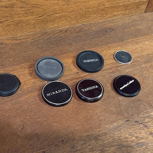 May include: A collection of black camera lens caps, some with brand names like "Yashica," "Miranda," and "Sony" printed in white. One small, silver-colored cap is also present. The caps are arranged on a wooden surface.