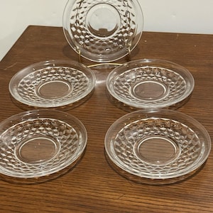 May include: A collection of five clear glass saucers featuring a diamond design. One saucer is presented on a gold-coloured stand. The remaining four saucers are arranged on a wooden surface. The saucers are circular with a central indentation.