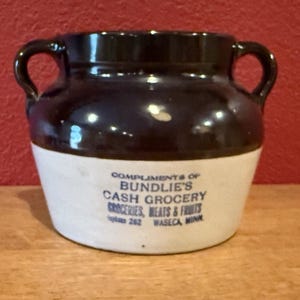 Puede incluir: Un tarro de cerámica antiguo con la parte superior marrón oscuro y dos asas. La parte inferior es blanca con texto azul que dice "Compliments of Bundlie's Cash Grocery". Una pieza clásica de menaje.