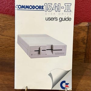 May include: A Commodore 1541-II Disk Drive user's guide. The cover features a light grey disk drive illustration. The text "COMMODORE 1541-II DISK DRIVE user's guide" is printed on the cover. The guide is displayed on a gold-coloured metal stand.