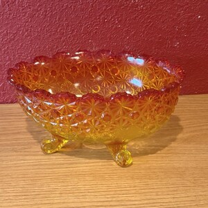 Könnte beinhalten: Eine bernsteinfarbene Glasschale mit Füßen und einem Sternmuster. Die Schale hat einen gewellten Rand und drei kleine Füße. Das Glas hat eine glänzende Oberfläche, und die Farbe geht von Gelb zu Orange über.