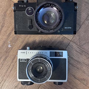 Könnte beinhalten: Zwei schwarze Vintage-Kameras mit silbernen Akzenten. Die obere Kamera hat die Aufschrift "T·L·S" und "ASA" sowie "SEARS" auf der unteren rechten Seite. Die untere Kamera ist mit "SEARS EASI-LOAD FC 600" beschriftet. Beide Kameras haben markante Objektive.