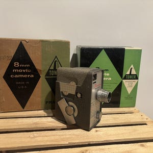 May include: Vintage 8mm movie camera with two boxes. The camera is dark gray and silver. The boxes are brown and green with black diamond and triangle designs. Text on the boxes reads "8mm movie camera" and "Made in USA."