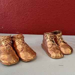 May include: Two pairs of metallic copper-colored baby shoes with lace-up fronts. Each shoe features a smooth, reflective finish, rounded toes, and visible laces tied at the top. The shoes are arranged in two pairs, showing a shiny, uniform texture.