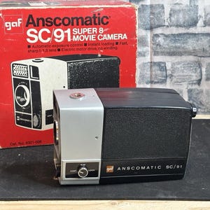 May include: A vintage GAF Anscomatic SC/91 Super 8 movie camera with its original box. The camera is black and silver. The box is red and white, with the camera model and features listed. The text "ANSCOMATIC SC/91" is on the front of the camera.