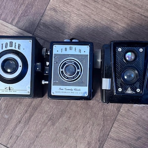 May include: Three vintage black cameras with silver and white accents are arranged on a wooden surface. The cameras are box-shaped, with the word "TOWER" prominently displayed on the front. One camera has the text "One-Twenty Flash" and "SEARS ROEBUCK AND CO. U.S.A."