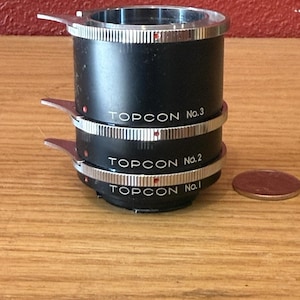 Può includere: Un set di adattatori per obiettivi per fotocamera Topcon neri, con tre pezzi cilindrici impilati con anelli argentati e il testo "TOPCON No. 1, 2, 3." Una piccola moneta è appoggiata accanto agli adattatori.