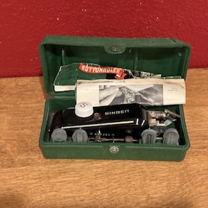 Puede incluir: Un accesorio de ojal Singer vintage en una caja verde. El ojal negro tiene una perilla blanca y está etiquetado como "SINGER". La caja contiene instrucciones y una etiqueta "BUTTONHOLER".