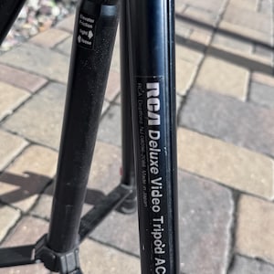 May include: Black tripod with a label that reads "RCA Deluxe Video Tripod AC031 RCA Deptford NJ 18096-2088 Made in Japan".