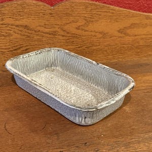 May include: A rectangular, grey and white speckled enamelware baking dish. The dish has a slightly textured surface and a rolled rim. The dish is on a wooden surface.