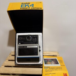Puede incluir: Una cámara instantánea Kodak EK4 vintage en su caja original, con la parte superior amarilla y el interior negro. La cámara es en blanco y negro con una franja azul. También hay un manual de Kodak, con impresiones en color y el texto "Kodak EK4 Instant camera".