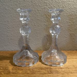 May include: Two clear glass candlesticks with intricate diamond and geometric patterns. The candlesticks have a flared base, a central orb-like design, and a top designed to hold a candle. They are displayed on a wooden surface.