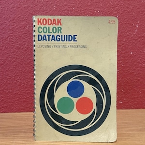 May include: A spiral-bound Kodak Color Dataguide manual with a tan cover. The text reads KODAK COLOR DATAGUIDE EXPOSING/PRINTING/PROCESSING and 4.95. A circular graphic features a black camera aperture design surrounding three colored circles.