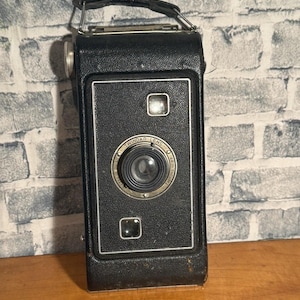 May include: A vintage black camera with a silver handle and trim. The camera has a square shape with a lens in the center and the words "TWINDAR LENS" around it. There are two small rectangular viewfinders above and below the lens.