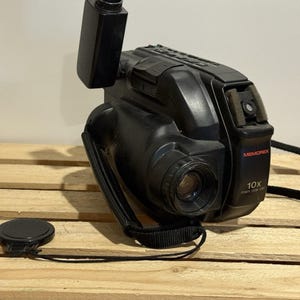 Puede incluir: Videocámara Memorex negra con lente, correa y tapa de lente. La videocámara tiene el texto "Memorex" y "10x Power Zoom Lens" impreso en el lateral. La videocámara está sobre una superficie de madera.