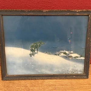 May include: Framed landscape art depicting two dark wolves on a snow-covered hill under a dark blue sky. The artwork is in a dark wooden frame, with a winter scene featuring small buildings and trees.