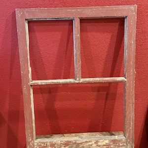 May include: A weathered, rectangular window frame with a divided pane design. The frame is painted a faded red with visible wear and tear, revealing hints of the underlying wood. The window frame is resting on a wooden surface against a red wall.