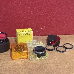 Puede incluir: Una colección de accesorios para cámara Soligor, que incluye una montura de lente, filtros y una funda. Los artículos están dispuestos sobre una superficie de madera contra un fondo rojo. La caja Soligor es amarilla con texto negro.
