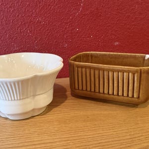 May include: A white ceramic bowl with a scalloped rim and ribbed detailing sits next to a brown rectangular planter with vertical grooves. Both are on a wooden surface against a red wall.