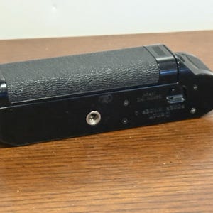Puede incluir: Un accesorio de cámara Canon Power Winder A negro. El accesorio tiene un agarre texturizado y un soporte de tornillo de metal. El producto es rectangular con bordes redondeados y un pequeño puerto en el lateral. El accesorio está sobre una superficie de madera.