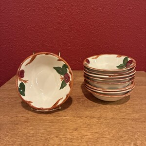 Puede incluir: Un juego de cuencos de cerámica blanca con borde marrón y diseño floral. Los cuencos tienen un interior blanco con un diseño pintado de hojas verdes y bayas rojas. Un cuenco está sobre un soporte metálico y los otros están apilados.