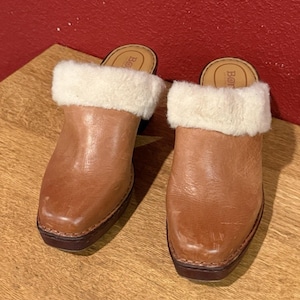 May include: A pair of brown leather clogs with a shearling cuff. The clogs have a square toe and a dark brown sole. The shearling cuff is cream-colored. The clogs are on a wooden surface.