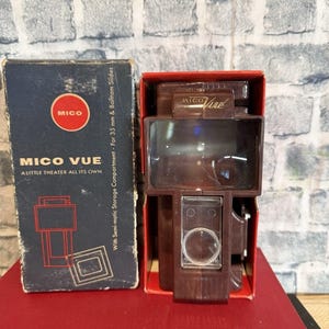 Könnte beinhalten: Ein Vintage Mico Vue Diabetrachter in seiner Originalverpackung. Die Schachtel ist dunkelblau mit roter und weißer Schrift und Grafiken. Der Betrachter ist braun und befindet sich in einer roten Schachtel. Der Text auf der Schachtel lautet "MICO VUE A LITTLE THEATER ALL ITS OWN."