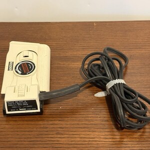 May include: Off-white electrical device with a black power cord. The device has a dial and a small rectangular label with text. The cord is bundled with a white plastic tie. The device is on a wooden surface.