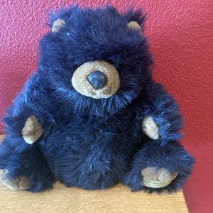 May include: A plush black bear toy with brown details on the ears, paws, and snout. The bear has a soft, furry texture and is sitting on a wooden surface. The bear's eyes are hidden by the fur.