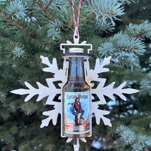 Pode incluir: Um enfeite de floco de neve de madeira com uma garrafa em miniatura de rum especiado Captain Morgan Original dentro. A garrafa é de vidro transparente com um rótulo marrom e uma tampa branca.