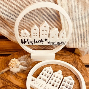 Könnte beinhalten: Ein weißes Holzschild mit einem runden Ausschnitt, der eine Silhouette von Häusern und den Text "Herzlich Willkommen" zeigt.