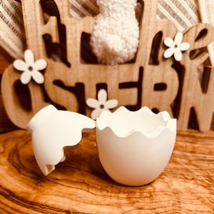 Könnte beinhalten: Ein weißer, eiförmiger Behälter aus Keramik ist in zwei Hälften zerbrochen und zeigt das Innere. Der Behälter steht auf einer Holzoberfläche.