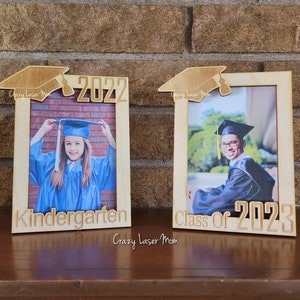 Puede incluir: Dos marcos de fotos de madera con gorros de graduación en la parte superior. El marco de la izquierda tiene una foto de una niña con una bata de graduación azul y dice "Kindergarten 2022". El marco de la derecha tiene una foto de un joven con una bata de graduación negra y dice "Class of 2023".