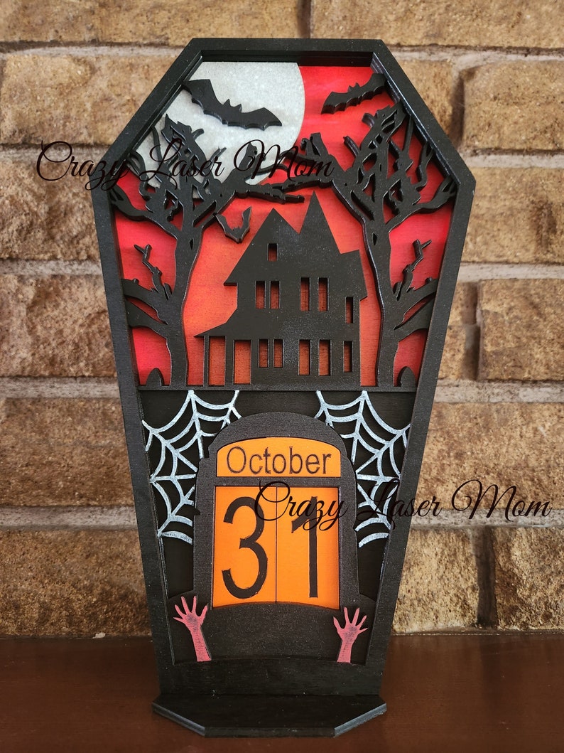 May include: Black wooden coffin-shaped Halloween countdown calendar with a haunted house, bats, and a full moon. The calendar features a spooky spiderweb and the date October 31st.