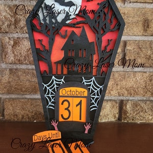 May include: A black wooden Halloween countdown calendar shaped like a coffin with a haunted house, bats, and a full moon. The calendar has a flip-up date display that reads "October 31". There are also two smaller flip-up numbers that read "01" and say "Days Until" on a separate piece of wood.