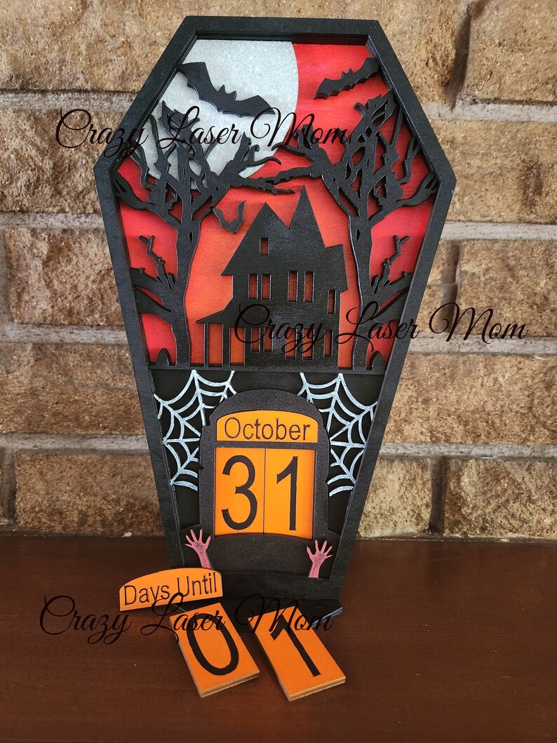 May include: A black wooden Halloween countdown calendar shaped like a coffin with a haunted house, bats, and a full moon. The calendar has a flip-up date display that reads "October 31". There are also two smaller flip-up numbers that read "01" and say "Days Until" on a separate piece of wood.