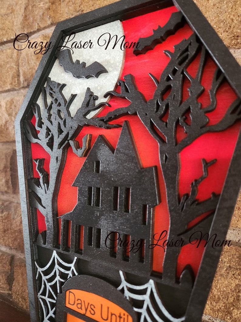 May include: A wooden Halloween countdown plaque with a black silhouette of a haunted house, trees, bats, and a moon. The plaque has a white spider web and the text "Days Until" on a black tombstone.
