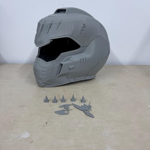 Puede incluir: Un casco gris impreso en 3D con un diseño detallado y bordes afilados. El casco tiene una visera y una superficie texturizada. Hay varias piezas pequeñas grises impresas en 3D dispersas alrededor del casco.