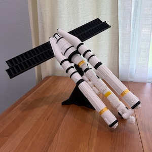 May include: A white and black model of a spacecraft with yellow accents. The model has multiple cylindrical components and a black rectangular wing-like structure. It is displayed on a wooden surface.