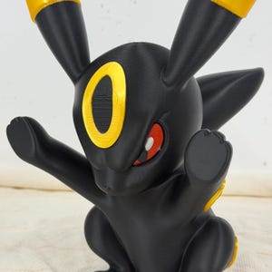 May include: A black figurine of a creature with long ears, featuring bright yellow accents on the ear tips and a yellow oval marking on the forehead. The creature has red eyes and is posed with both arms raised in a forward-facing stance.