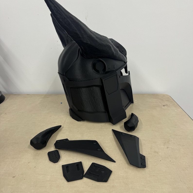 Puede incluir: Un casco de pl&aacute;stico negro con un gran cuerno en la parte superior. El casco est&aacute; en piezas, con el cuerno, la visera y las piezas laterales separadas.