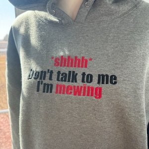 Könnte beinhalten: Grauer Kapuzenpullover mit dem Text "*shhh* don't talk to me I'm mewing" in schwarzer und roter Stickerei.