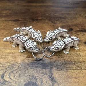 Puede incluir: Cuatro llaveros de gecko marrones y blancos con anillos de llaves plateados. Los geckos están dispuestos en círculo, mirando hacia adentro.