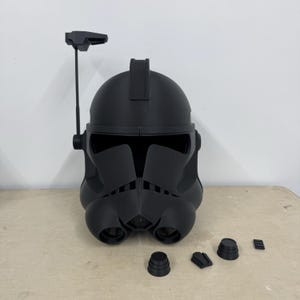 Puede incluir: Un casco de plástico negro con una cresta elevada en la parte superior y una pequeña antena. El casco tiene una visera y una máscara facial con una serie de barras verticales. Hay cuatro pequeñas piezas de plástico negro en la mesa delante del casco.