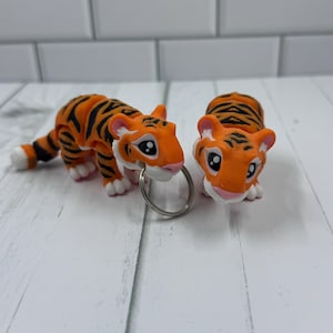 Puede incluir: Dos figuritas de llavero de tigre naranja y negro con caras blancas y ojos negros. El tigre de la izquierda sostiene un llavero plateado.