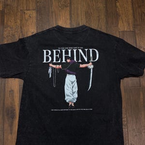 Pode incluir: T-shirt preta com a palavra "BEHIND" em branco. A camisa apresenta uma ilustração de uma pessoa com correntes e uma espada. O texto "TO THE ONE WHO LEFT IT ALL" e "THEY WOULD ALL BEAR WITNESS TO THE BARE FLESH OF THE ONE WHO IS FREE" também estão impressos.