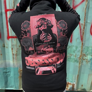 Puede incluir: Una sudadera con capucha negra que presenta un gran gráfico rojo en la espalda. El diseño incluye a una persona sosteniendo un jarrón de rosas, el texto "BE CAREFUL WHO YOU WISH FOR", la palabra "OBSESSION" en negrita y una imagen pequeña.